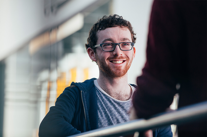 smiling male student wearing glasses facing someone with their back to camera