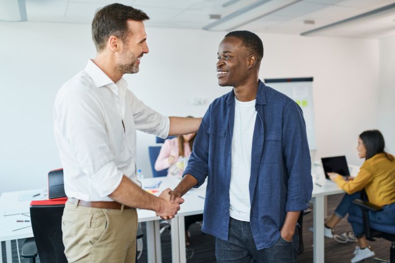 White dark haired gentleman wearing a white shirt and beige chino trousers on the left shaking hands with a black  dark haired gentleman wearing a navy denim shirt and dark coloured trousers on the right.