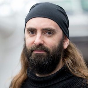 image of Gareth Hughes, a Caucasian male with long dark brown hair and beard wearing a black top and black cap