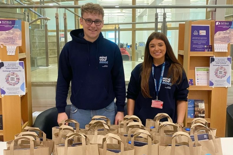 Image shows two previous Mind Your Mood Coordinators distributing self-care packs to students in the McClay Library.