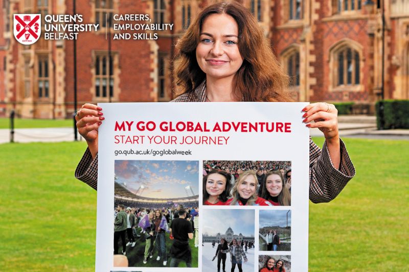 female student standing outside Queen's university main building holding up a cardboard post about go global week