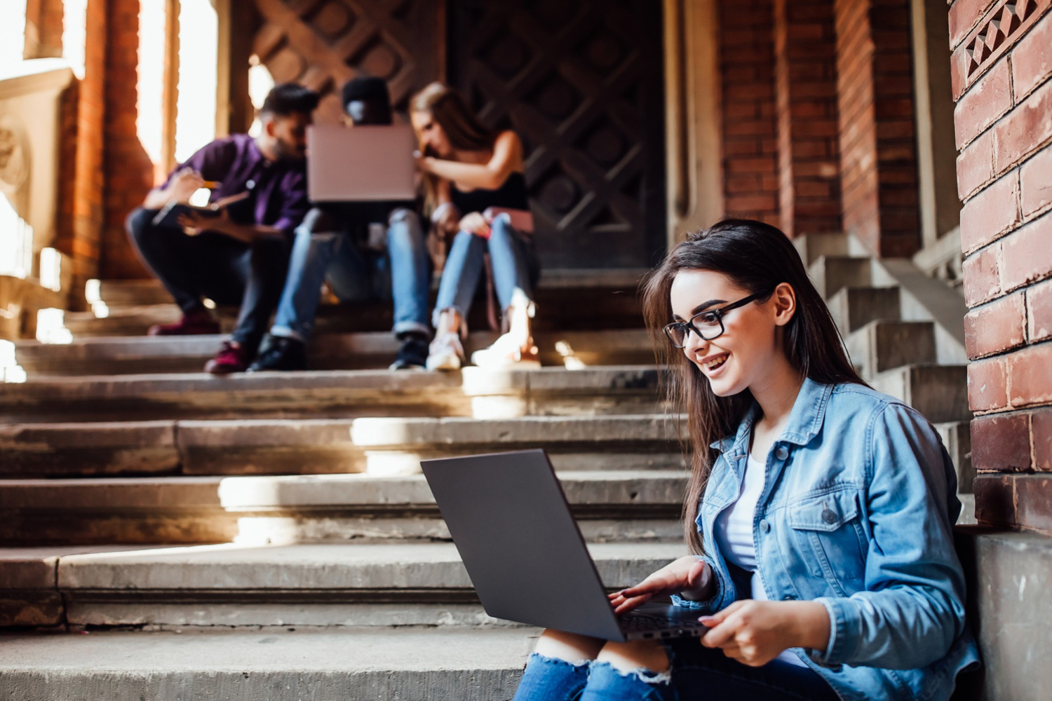 Female student working on a laptop, sitting on stairs with other students sitting in the background