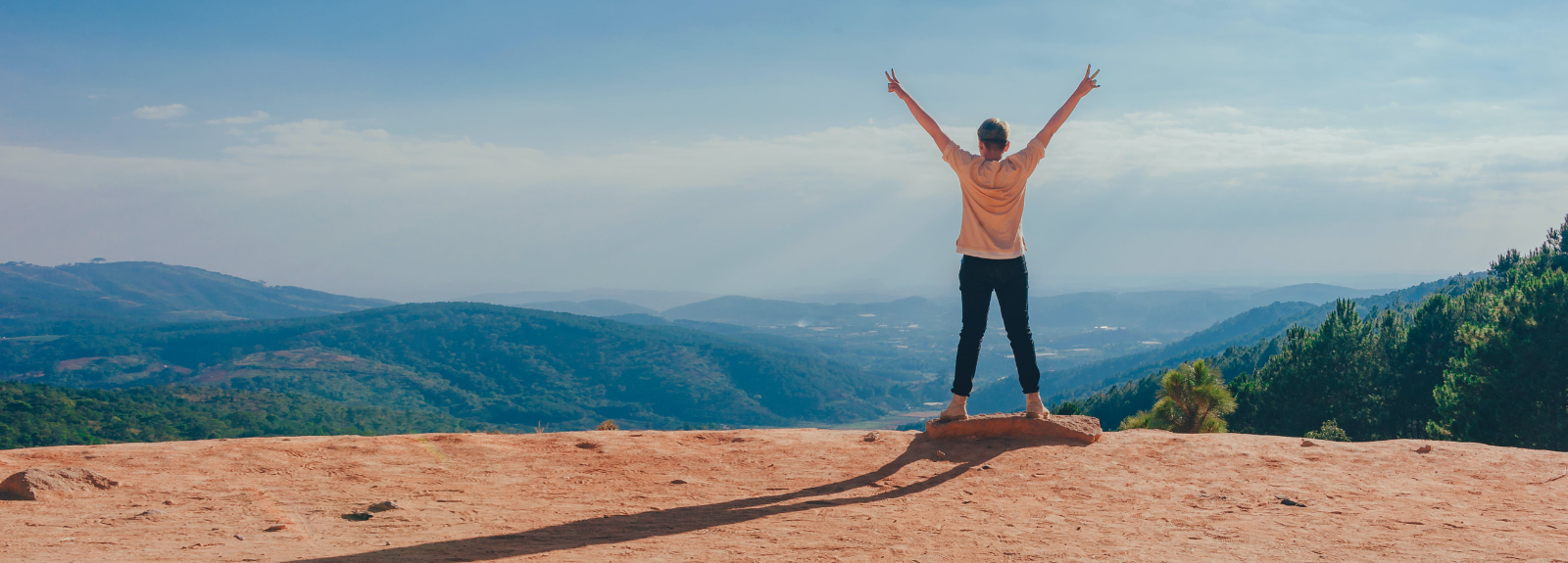 male standing on a sunny cliff top over looking sea with arms in the air