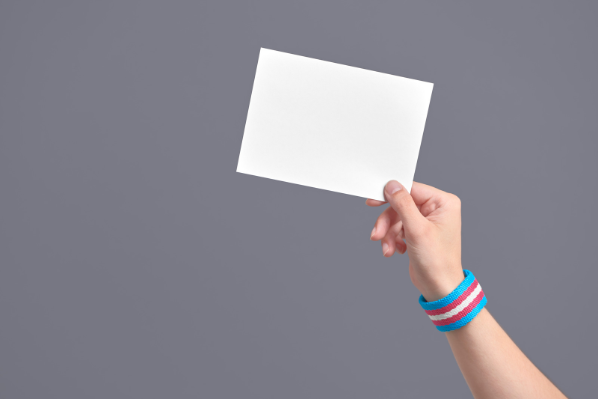 Image shows a hand holding up a white card. The person is wearing a wristband in the colours of the transgender flag.