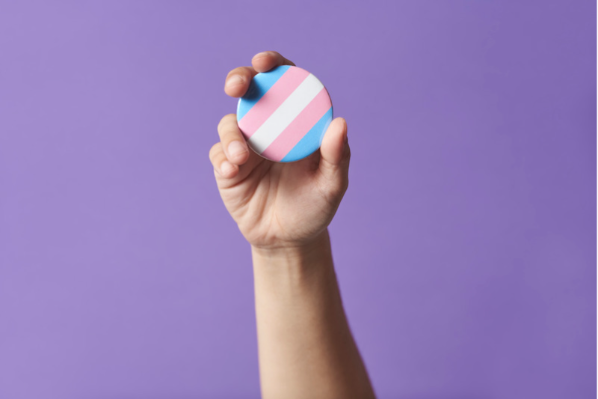 Image shows a hand holding a circular transgender flag. The background is purple.