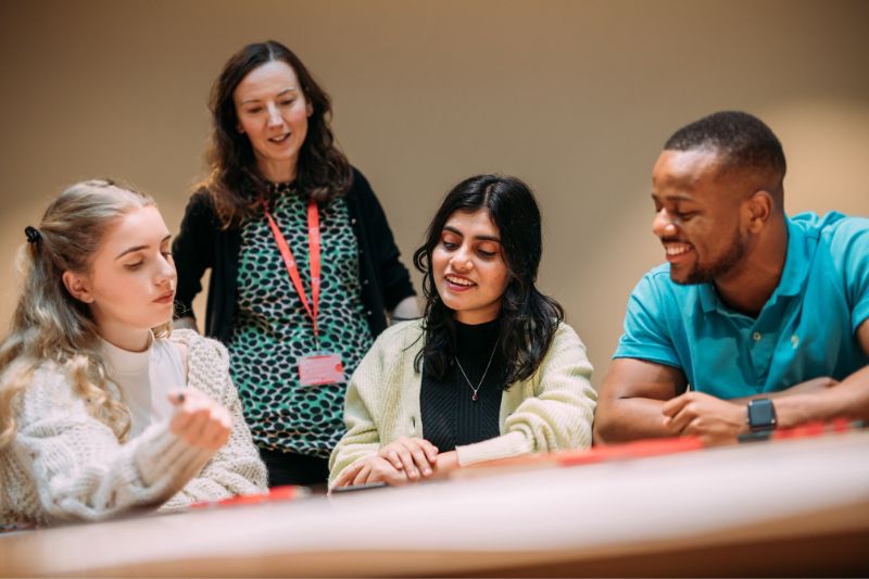 three students sitting at a table talking whilst a staff member listens to their conversation