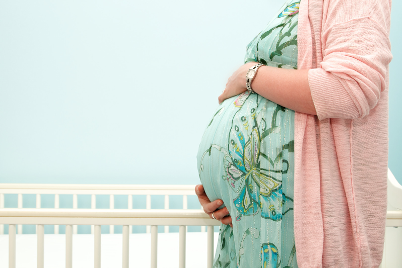 Image shows a pregnant woman holding her bump in front of a cot