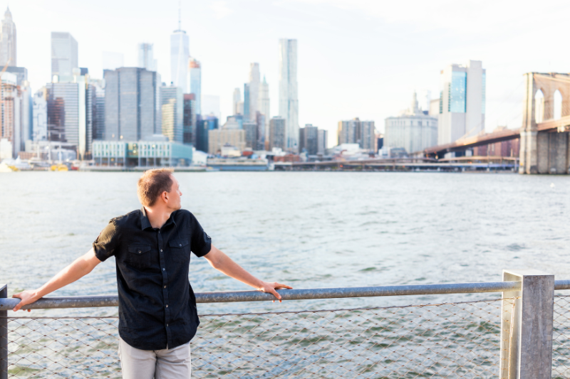 male student standing at a barrier of a river looking at a cityscape