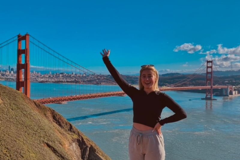 female student standing with an arm in the air at a scenic spot overlooking a coastal scene