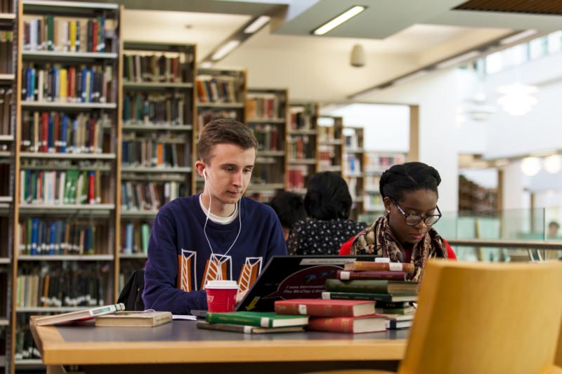 two students sitting at a desk in the library working on laptops with textbooks in front of them and bookcases in the background