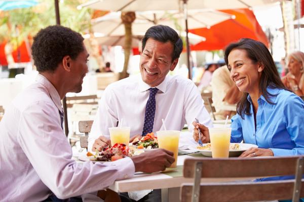 Three people in business attire, sitting outside around a table eating lunch under parasols, with three glasses of orange smoothies or juice.  There is one dark haired younger man facing an older Asian looking man and a dark haired lady in a blue long sleeved blouse