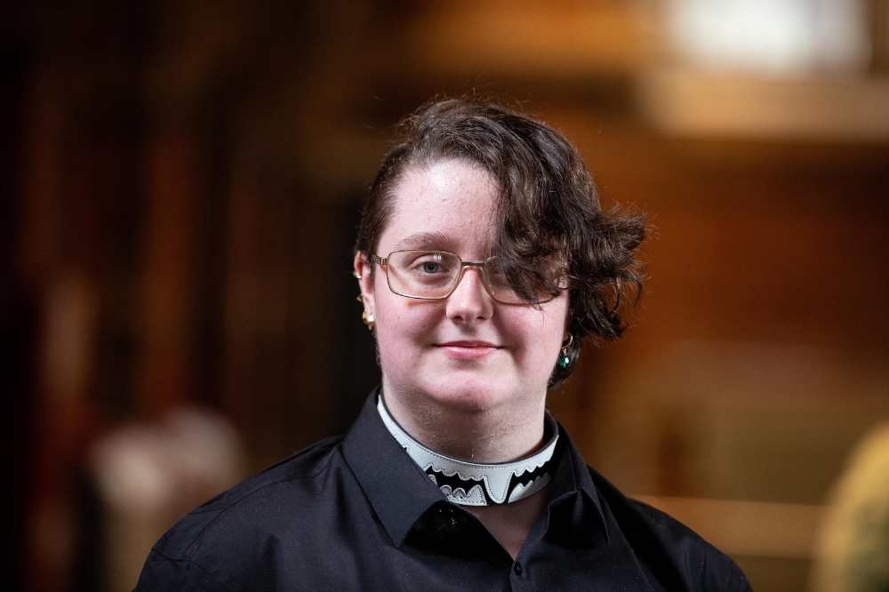 A person with short, curly hair and glasses stands indoors, wearing a black shirt with a clerical-style collar featuring a decorative black and white design.