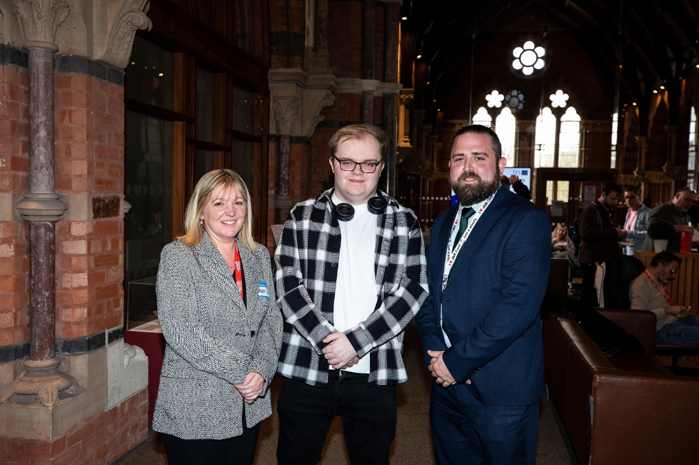 Three people standing together indoors in front of a brick and stone wall at Queen’s University Belfast. The woman on the left is wearing a grey blazer and smiling. The young man in the centre is wearing glasses, headphones around his neck, and a black-and-white checkered shirt. The man on the right is dressed in a navy suit.