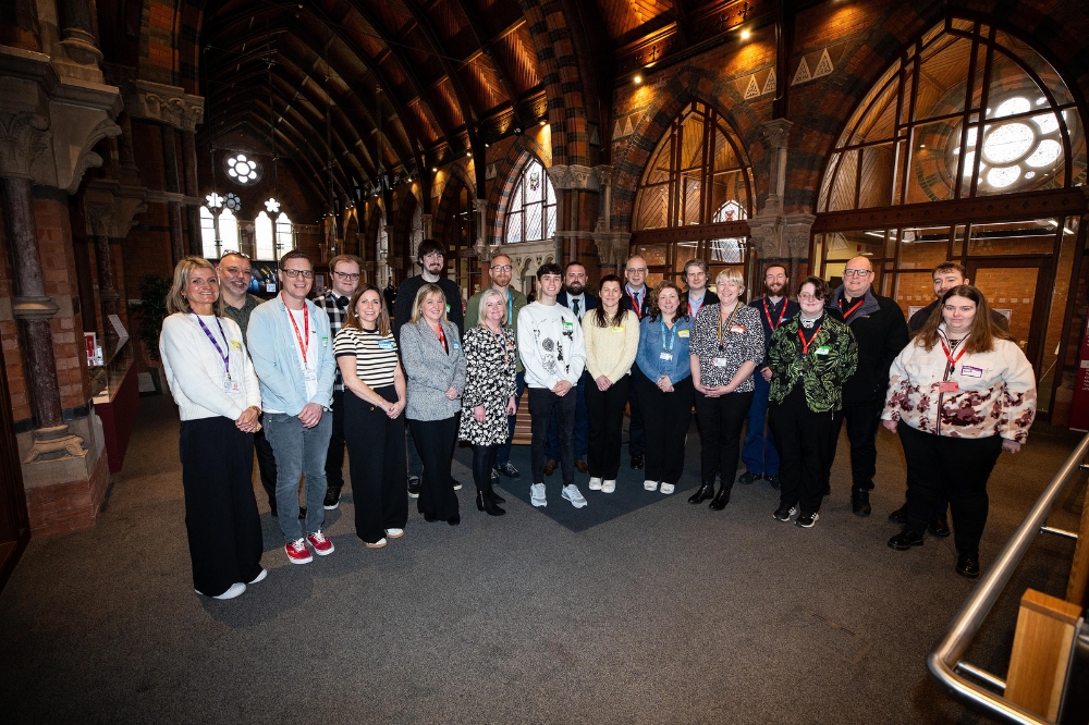 Group photo of attendees at the Inclusive Employment Scheme catch-up event, taken inside the Lanyon Building at Queen’s University Belfast. The group includes participants, Queen’s staff, and representatives from external organisations, standing together and smiling for the camera in a warmly lit, arched hallway.