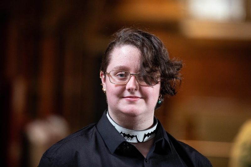 A person with short, curly hair and glasses stands indoors, wearing a black shirt with a clerical-style collar featuring a decorative black and white design.
