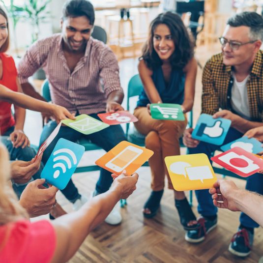 A diverse group of people sit in a circle holding colourful cards with communication and social media icons, taking part in a group discussion or workshop.