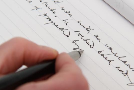 A Hand holding a pen writing with black ink on a white sheet of paper