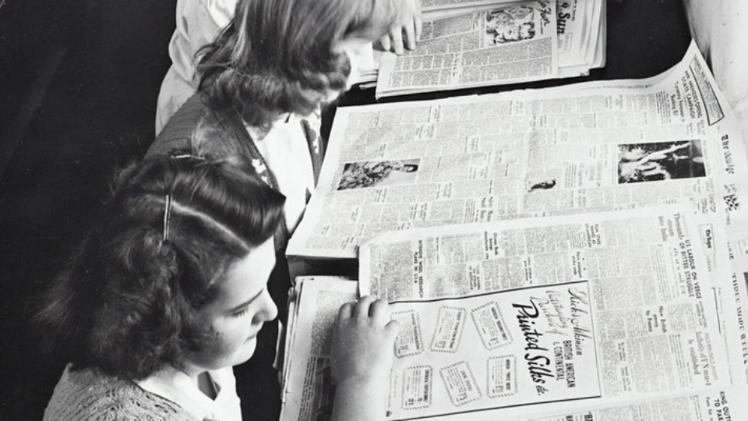 black and white image of young girls looking at newspaper archives in the early to mid 20th century