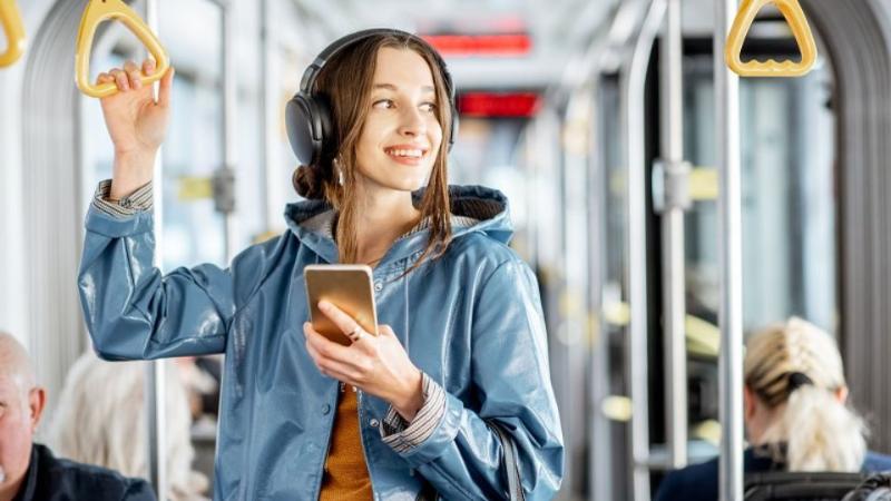female student on a bus standing in bus aisle with earphones in looking at phone