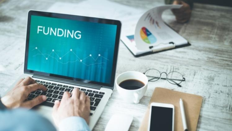 Person typing on laptop with 'Funding' on the screen, coffee and a mobile phone next to it
