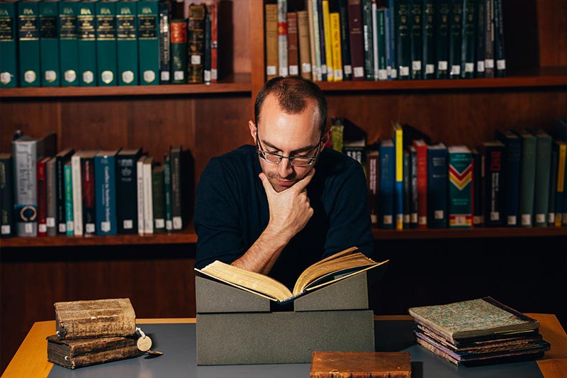 male student with glasses reading an old textbook whilst sitting in front of a bookcase