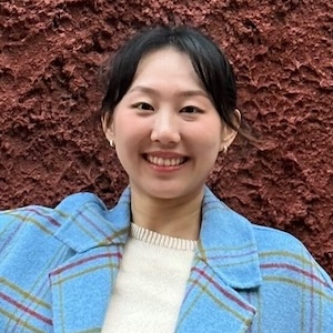 Portrait of Junyi Wang, smiling, in blue coat in front of red wall