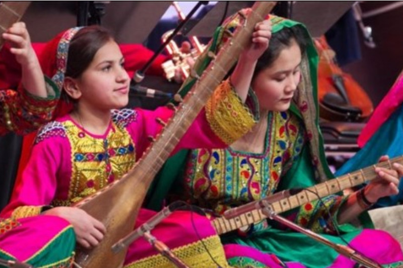 The image shows two young Afghan women in traditional clothing, playing stringed instruments as part of an Orchestra