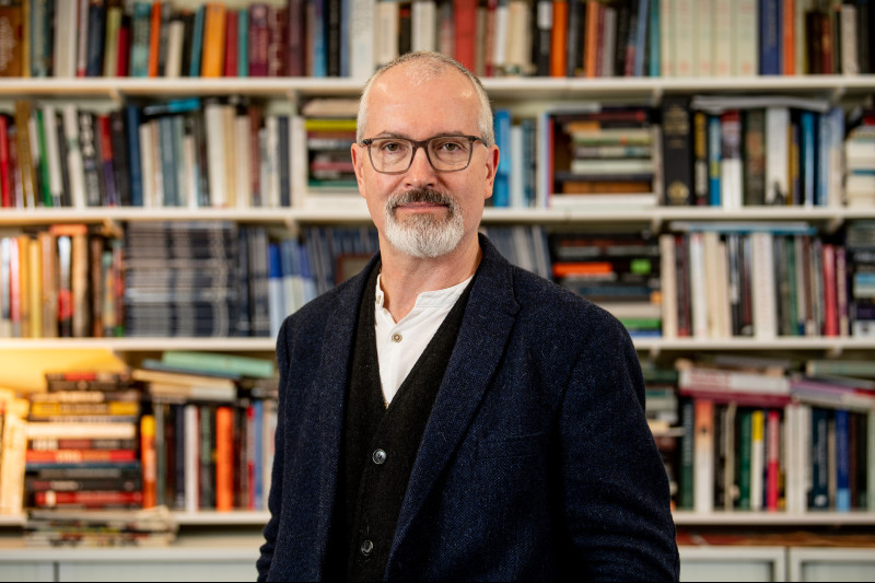 The photograph is a headshot of Professor Richard English in front of a wall of books on shelves.
