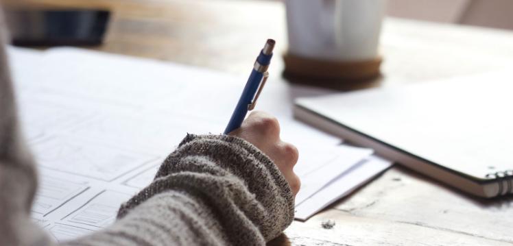 Hand writing at a desk with a cup in the background