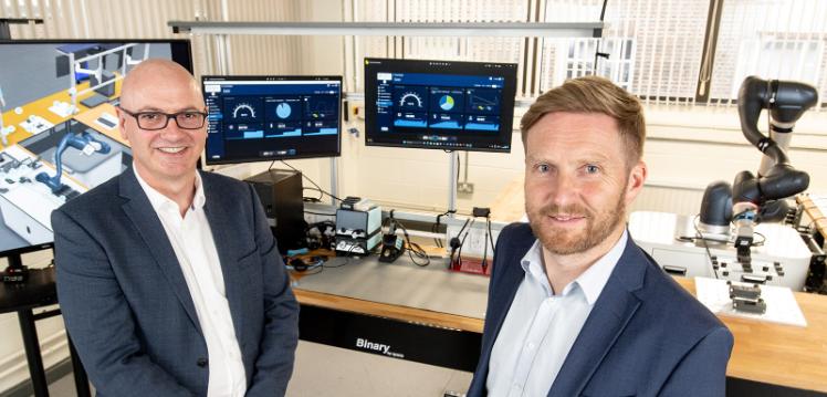 Two men stand in front of computer screens and robots
