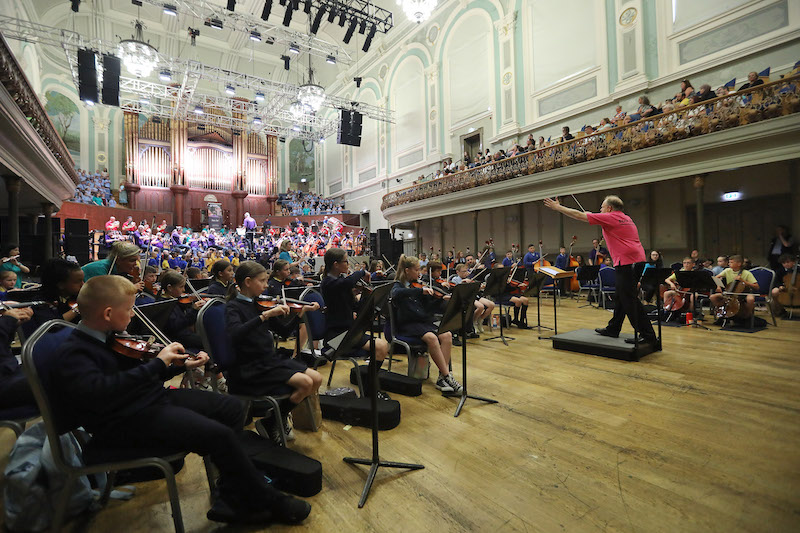 Crescendo concert 2023; conductor gestures to the Ulster Orchestra and school children playing in Ulster Hall