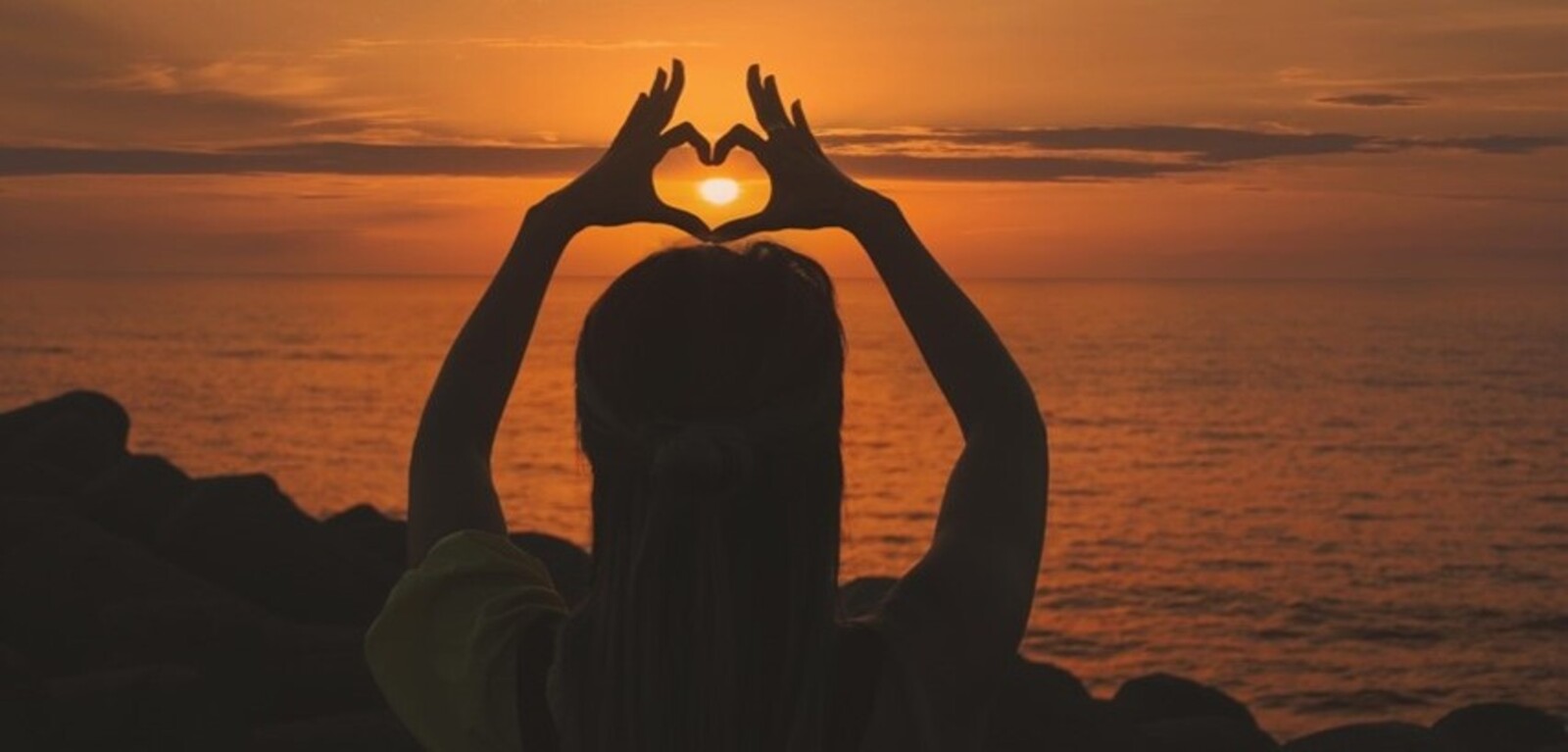 Person making a heart with their hands in front of sunset