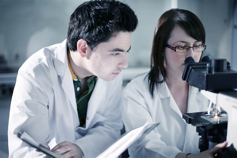 female student examines slides on a microscope whilst a male student takes notes