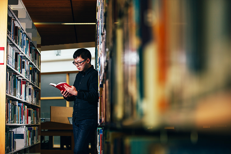 male student leaning against a bookshelf and reading a book