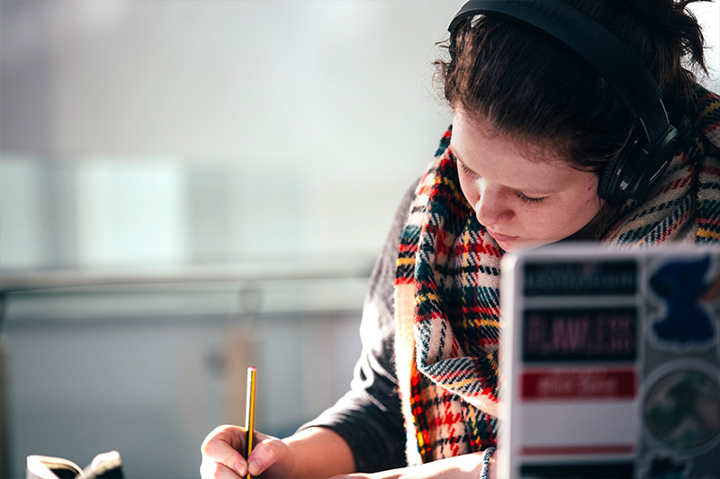 close up of a Female student wearing headphones and taking notes whilst using a laptop
