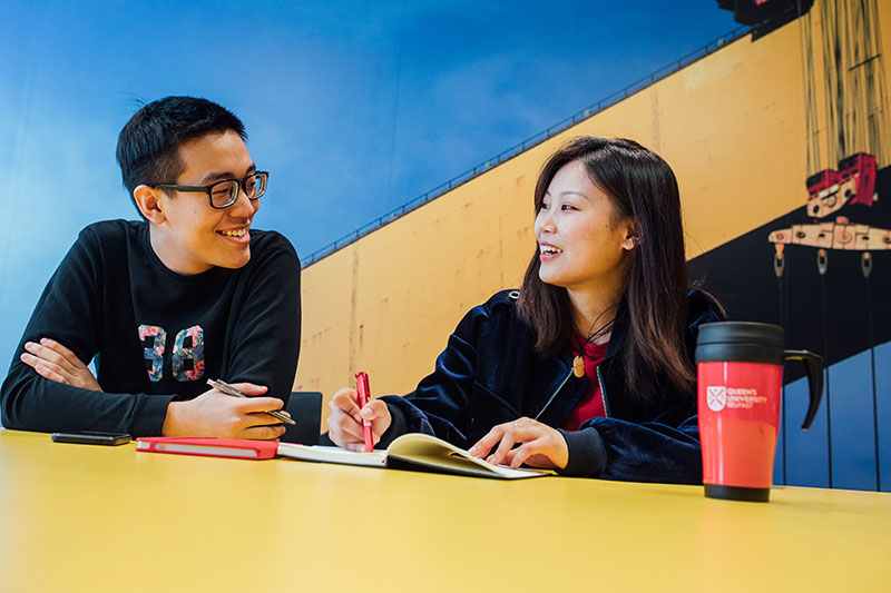 Two students seated in front of a feature wall depicting the iconic Harland and Wolff cranes