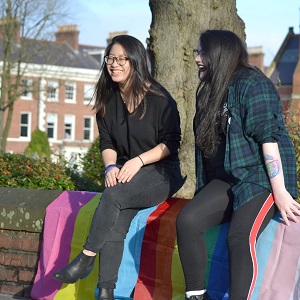 Two female students sit on a wall draped with LGBT flags and Trans rights flags