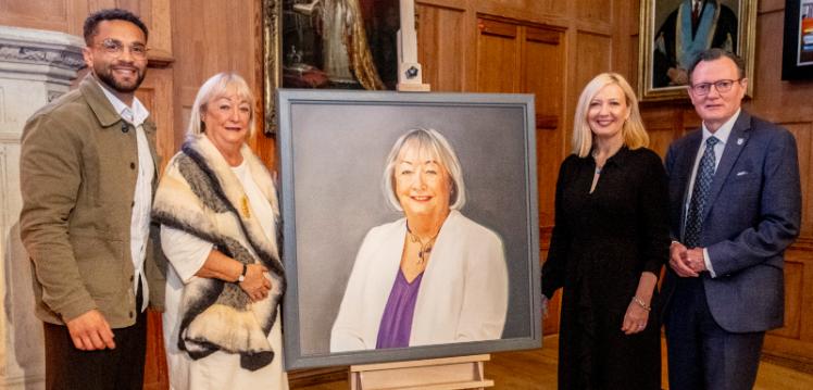 Two men and two women stand either side of a large portrait in a University's grand hall