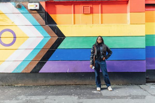 Student Natasha stands in front of inclusion flag wall art