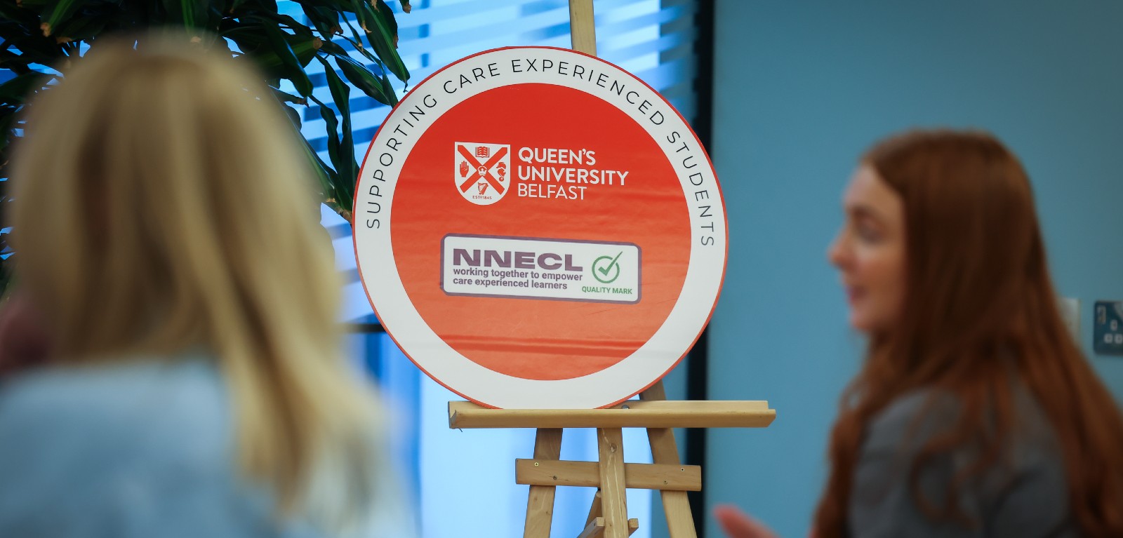 A university award sits in focus on an easel, two students sit in the foreground out of focus