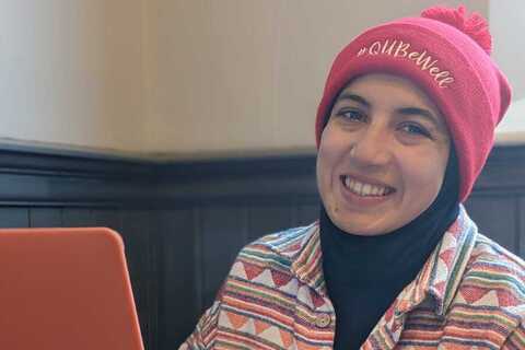 Student smiling in front of laptop while wearing QUBeWell hat