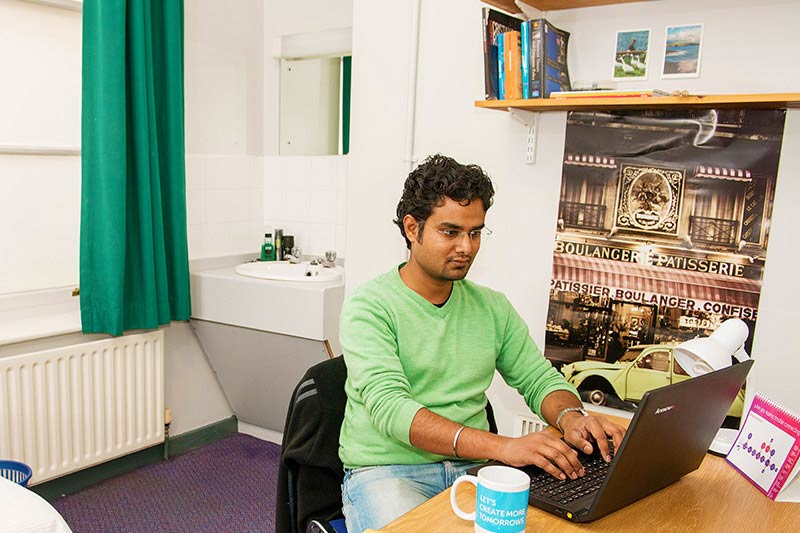 a male student sitting at the desk in his room and working on his laptop 