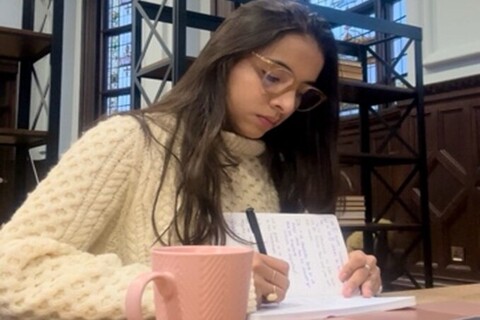 Student writing in notebook with a mug an laptop in front of them