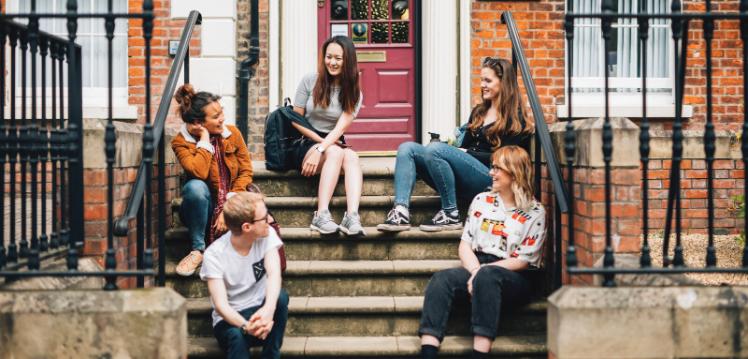 Group of five students laughing together while sitting on steps of Queen's campus building