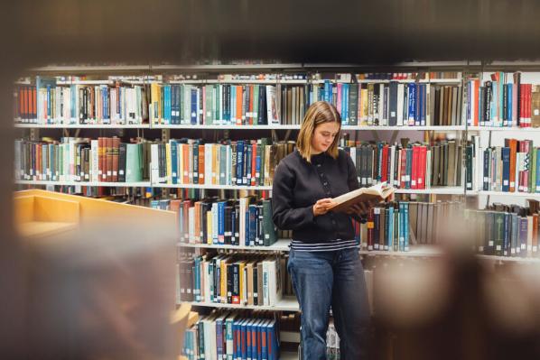 Student Tessa reads book beside library bookcase