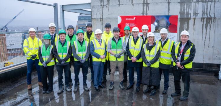 A large group of people wearing high-visibility jackets and hard hats stand on the top floor of a tall building being constructed. The background shows grey clouds and Belfast city