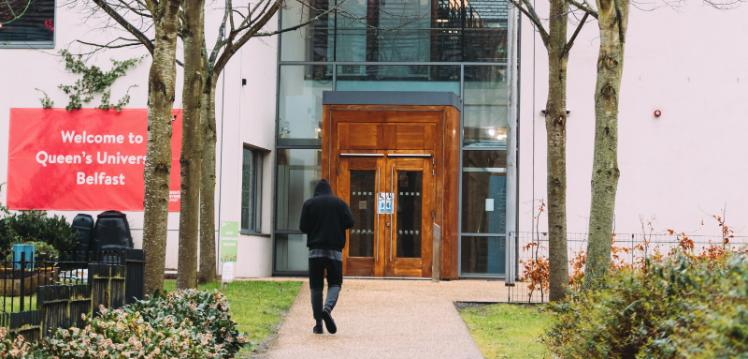 Student walking towards building with 'Welcome to Queen's University Belfast' sign on the left