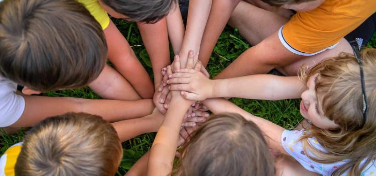 A group of children sitting in a circle outdoors placing their hands together in the centre as a symbol of teamwork and unity.