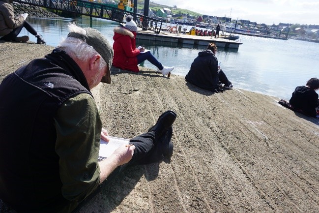 older man writing on piece of paper by the water