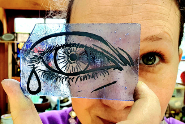 woman holds up piece of glass with drawn on eye